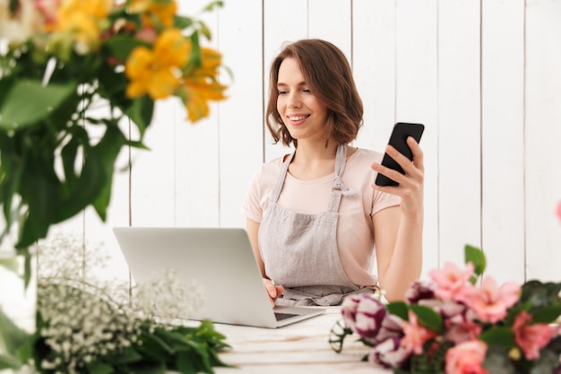 Cute cheerful florist lady using laptop computer chatting by mobile phone.