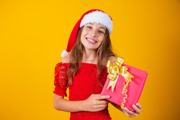 Cute cheerful girl in a Christmas hat on a colored background holding a gift