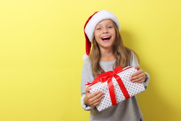 Cute cheerful girl in a christmas hat on a colored background holding a gift