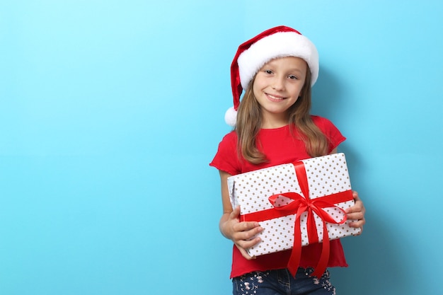 Cute cheerful girl in a christmas hat on a colored background holding a gift