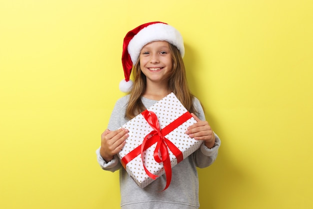 Cute cheerful girl in a christmas hat on a colored background holding a gift