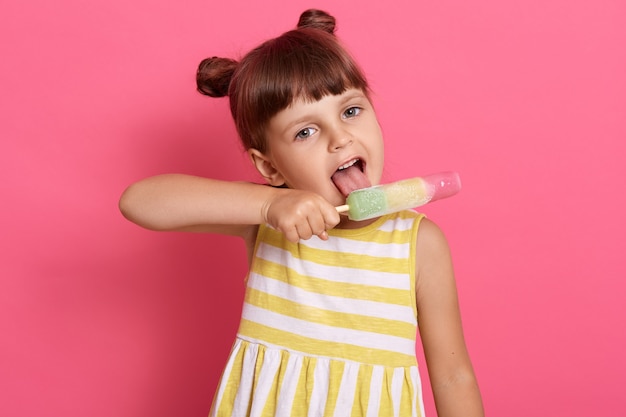 Cute child girl holds ice cream in hand and keeps mouth opened, licking sorbet, funny toddler child posing isolated over pink background, charming little lady with knots.