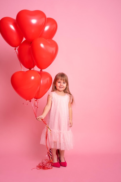 Cute child girl in pink dress with red heart balloons on pink background valentine day