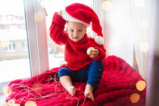 Cute child sitting near the window in red Santa hat. Celebrating Christmas at home