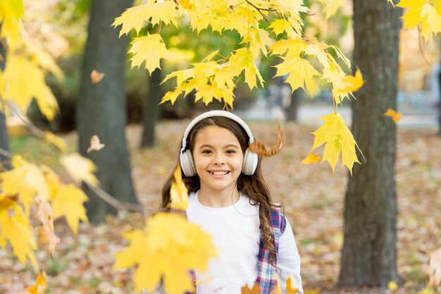 Cute child smile with stereo headphones. Melody of autumn. Schoolgirl listening modern headphones. Headphones technology. Falling leaves. Happy little girl wear headphones autumn nature background.