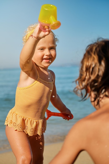 Cute child in yellow swimsuit having fun with watering can and looking at boy near sea on sandy beach