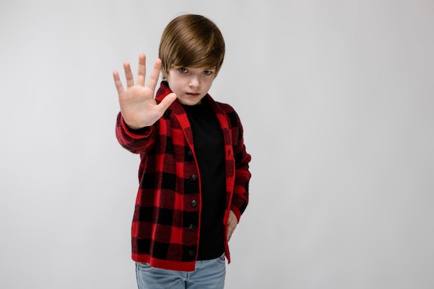 Cute confident sad little caucasian boy in checkered shirt showing stop sign on grey 