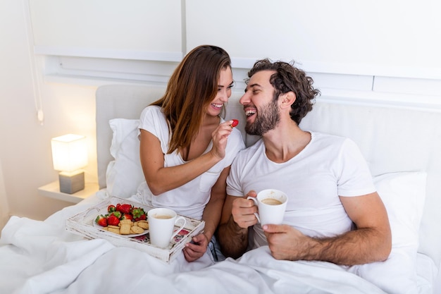 Cute couple having breakfast in bed in the bedroom. Beautiful woman feeding her boyfriend strawberries in bed while having breakfast and coffee in bedroom