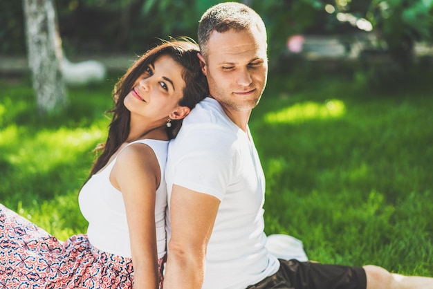 Cute couple in love sitting on a picnic blanket outdoor Caucasian happy dreamy man and smiling brunette woman sitting on green grass in park