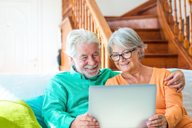 Cute couple of seniors watching movies having fun using laptop at home sitting on the sofa together Two mature pensioners holding a computer pc smiling indoor