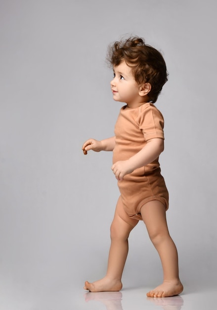 Cute curly baby in a beige bodysuit makes his first steps looks up with interest on a gray background side view