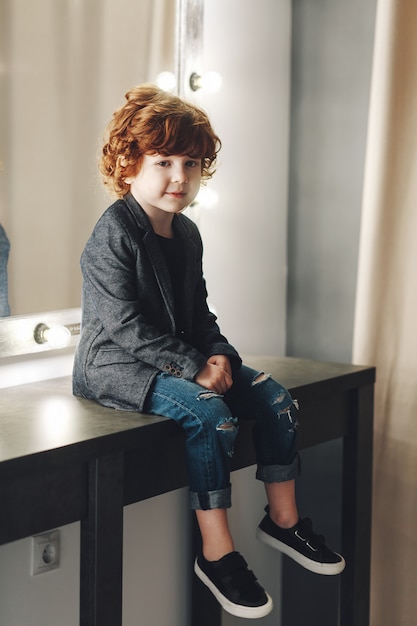 Cute curly boy child in jacket and ripped jeans posing near the mirror and smiling