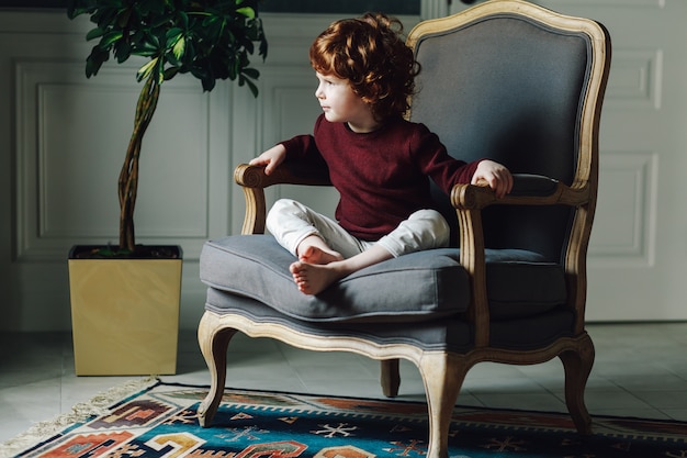 Cute curly boy sitting relaxed in comfortable armchair and looking away