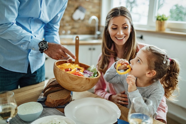 Cute daughter and her parents having lunch in dining room. Focus is on little girl eating bell pepper.