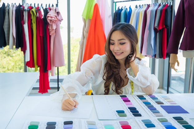 Cute and enerfyful female Asian designer working at desk in design room among clothes and fabric color sample.