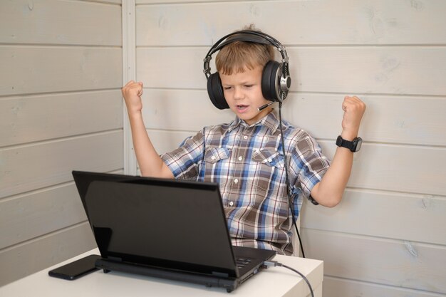 Cute European boy in headphones with a microphone happily throws his hands up sitting on a laptop.