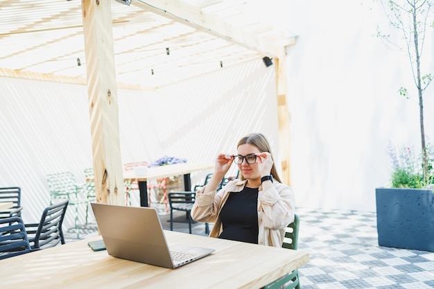 Cute european woman sitting at outdoor cafe table with laptop and cup of coffee smiling woman in glasses enjoying telecommuting in cafe or studying online