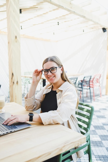 Cute european woman sitting at outdoor cafe table with laptop and cup of coffee smiling woman in glasses enjoying telecommuting in cafe or studying online