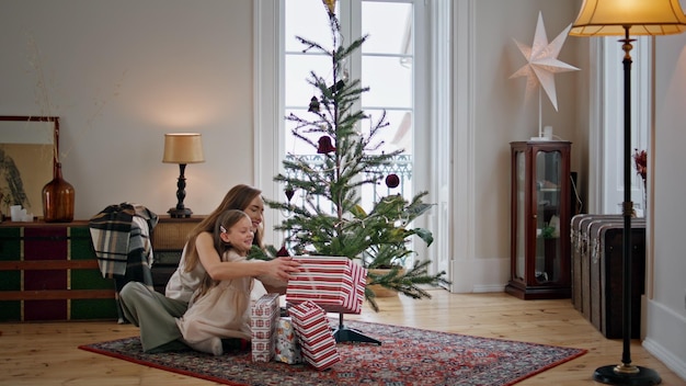 Cute family shaking gift box near xmas tree Woman embracing girl at cozy home