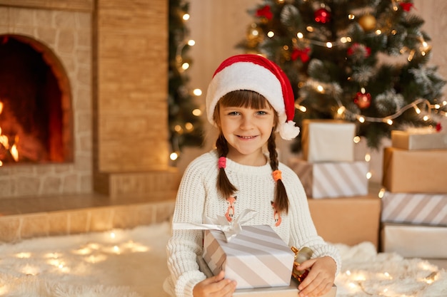 Cute female child in red hat on floor at home. Christmas celebration with presents, little girl posing with gift boxes,  dresses white jumper, sits near fireplace and xmas tree