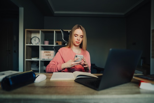 Cute female student sitting at home at desk with laptop and books and uses smartphone