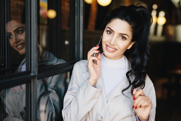 Cute female with wavy dark hair charming eyes wellshaped lips and white even teeth wearing white clothes having red manicure communicating over telephone touching her hair with her gentle hand