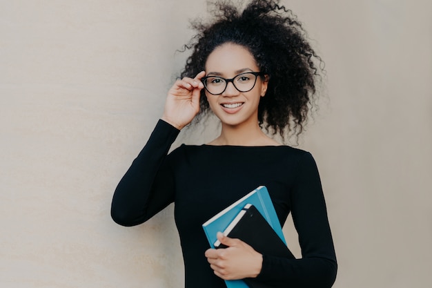 Cute frizzy young woman with gentle smile, keeps hand on rim of glasses, wears casual black jumper, holds notepad