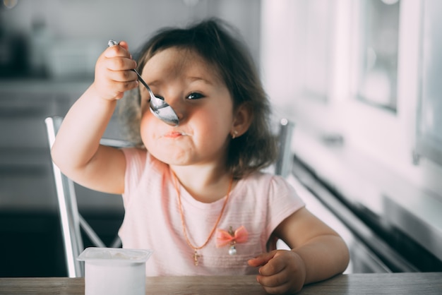 Cute, funny baby girl eating yogurt in the kitchen in a pink dress in the afternoon cute