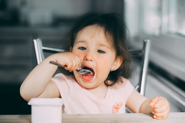 Cute, funny baby girl eating yogurt in the kitchen in a pink dress in the afternoon cute