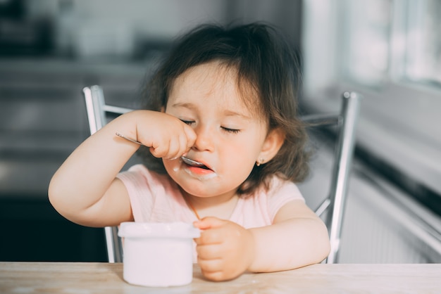 Cute, funny baby girl eating yogurt in the kitchen in a pink dress in the afternoon cute