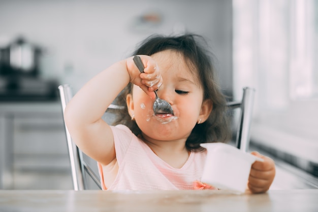 Cute, funny baby girl eating yogurt in the kitchen in a pink dress in the afternoon cute