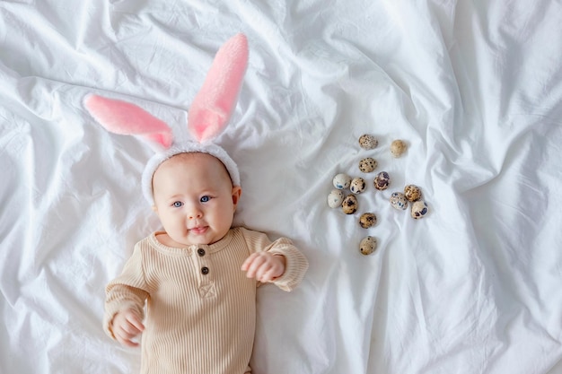 A cute funny baby with bunny ears costume dressed up for Easter laying on white bed linen sheet