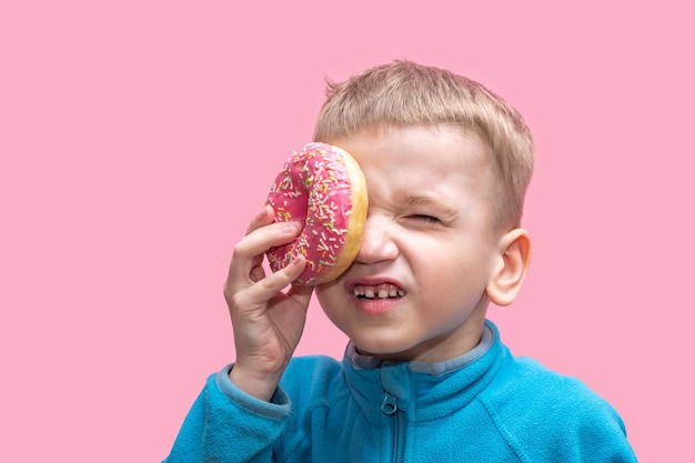 A cute funny boy in a blue sweater holds a bright pink donut near his eye and squints on a pink background Cheerful childhood concept Sweet food for kids concept