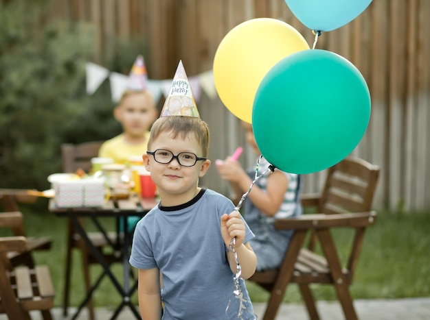 Cute funny four year old boy celebrating his birthday with family or friends in a backyard Birthday party Kid wearing party hat and holding balloon in a hand