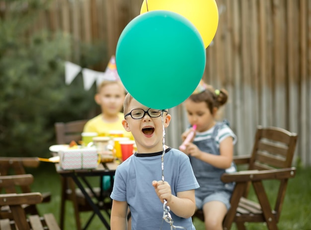 Cute funny four year old boy celebrating his birthday with family or friends in a backyard Birthday party Kid wearing party hat and holding balloon in a hand