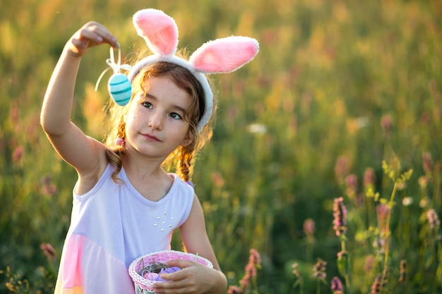 Cute funny girl with painted Easter eggs in spring in nature in a field with golden sunlight and flowers. Easter holiday, Easter bunny with ears, colorful eggs in a basket. Lifestyle