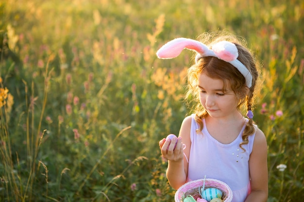 Cute funny girl with painted Easter eggs in spring in nature in a field with golden sunlight and flowers Easter holiday Easter bunny with ears colorful eggs in a basket Lifestyle