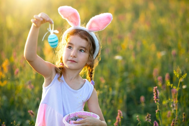 Cute funny girl with painted Easter eggs in spring in nature in a field with golden sunlight and flowers Easter holiday Easter bunny with ears colorful eggs in a basket Lifestyle