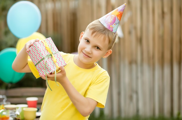 Cute funny nine year old boy celebrating his birthday with family or friends in a backyard Birthday party Kid wearing party hat and holding gift box