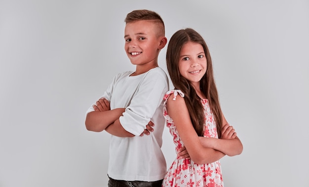 Cute girl and boy on a gray background are back to each other, arms crossed and smiling.