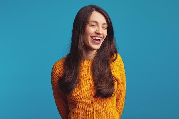 Cute girl in casual orange jumper smiling broadly with closed eyes against