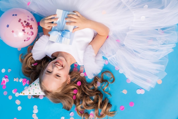 Cute girl in a fluffy white dress with a gift in her hands lies on a blue background in confetti