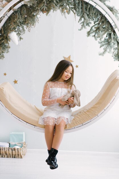 Cute girl holding a soft favorite toy and sitting on a swing decorated for Christmas