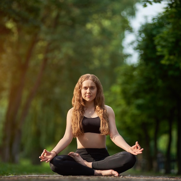 Cute girl is meditating in lotus position Padmasana Asana on the background of blurred green park