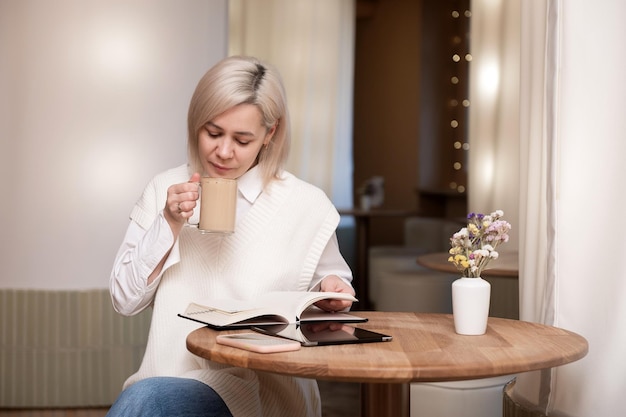 A cute girl is sitting with a tablet and a diary in a cafe and drinking coffee