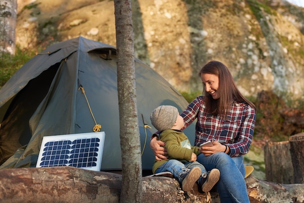 Cute girl looks at child sitting on log in forest near solar pan