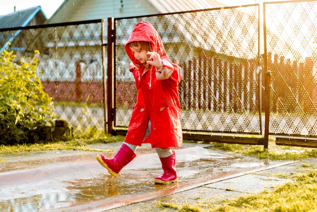 Photo cute girl in red jacket is jumping in puddle.the setting warm summer or autumn sun. summer in village.