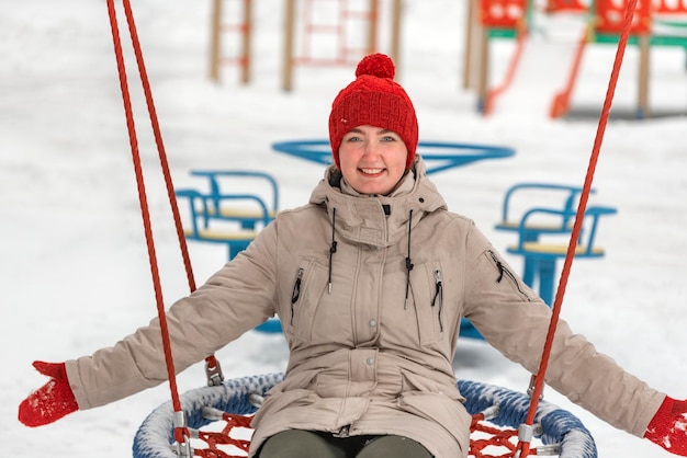 Cute girl in red knitted hat and mittens cheerfully rides on swing on playground Young woman has fun in winter outdoors