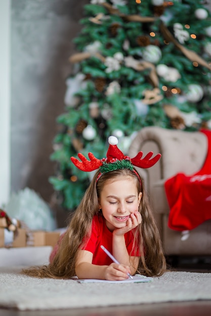 A cute girl in a Santa hat writes a letter to Santa near the Christmas tree. Happy childhood, a time for fulfilling desires.