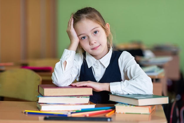 A cute girl schoolgirl sits at a desk surrounded by books Looks into the camera School life